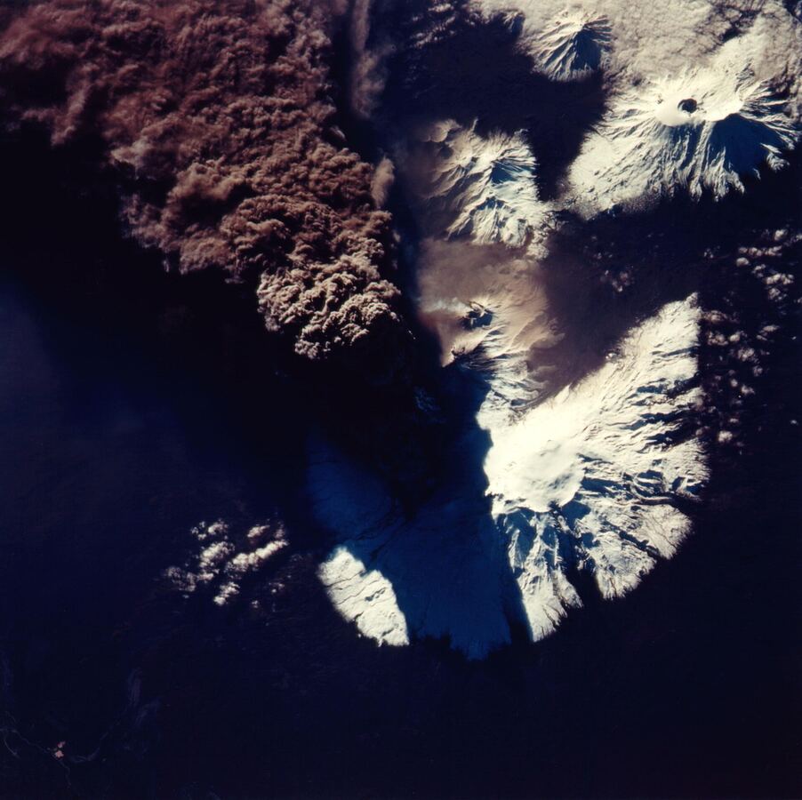Vista orbital de vulcões cobertos de neve em Kamchatka, com uma pluma marrom de cinza se espalhando a partir do Kliuchevskoi