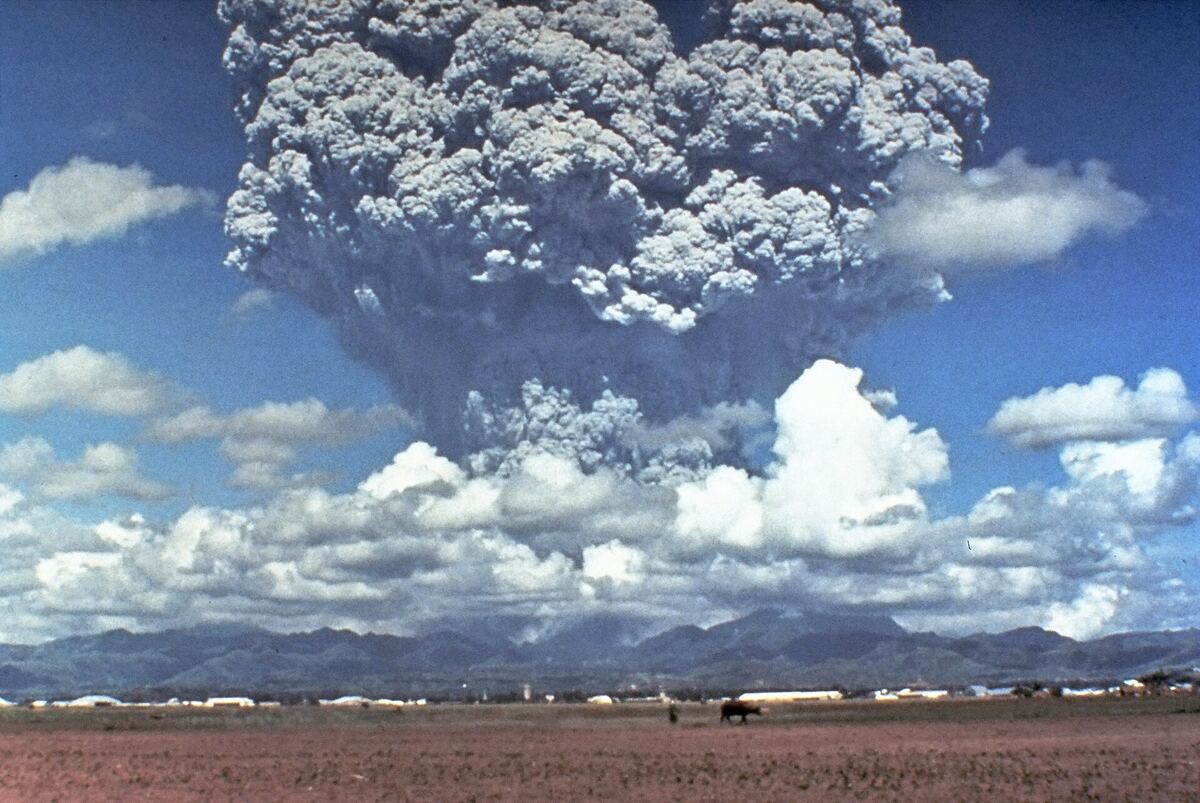 Uma enorme coluna eruptiva cinza subindo acima de nuvens e montanhas, vista através de um campo plano a partir da base aérea de Clark